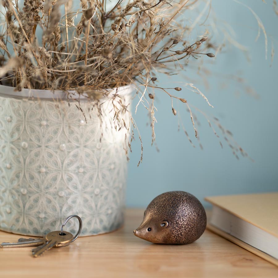 Small Bronze hedgehog sculpture on a wooden top next to a set of keys and a vase with dried flowers and book