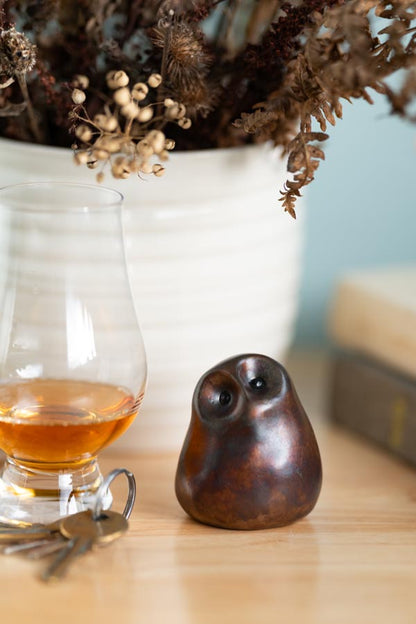 Brown coloured small bronze owl sculpture on a wooden surface next to some keys and a small glass with a white vase and dried flowers in the background
