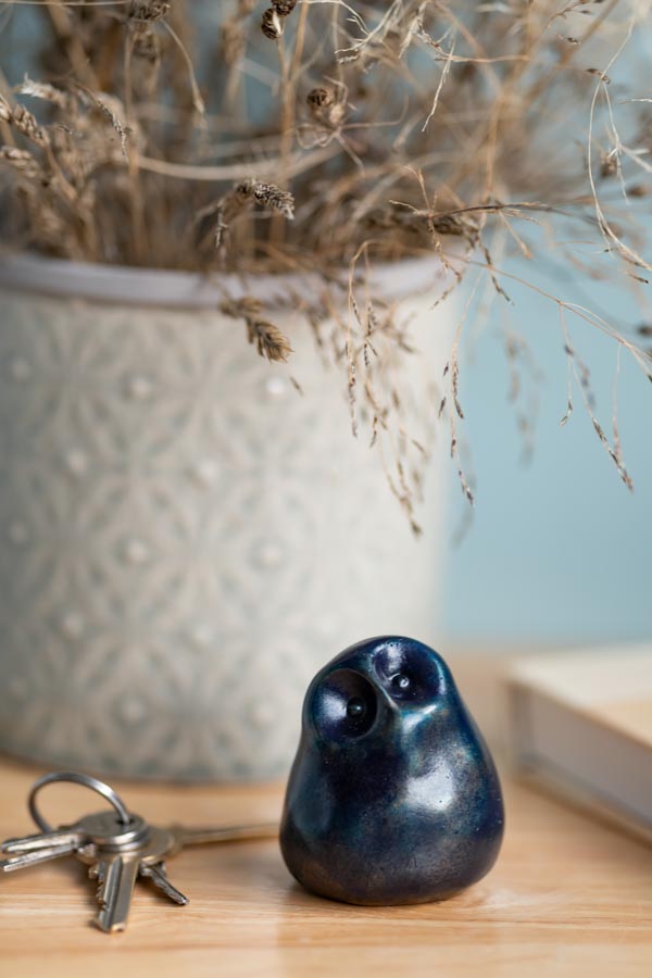 Deep blue small bronze owl sculpture on a wooden surface next to some keys with a vase in the background with dried grass