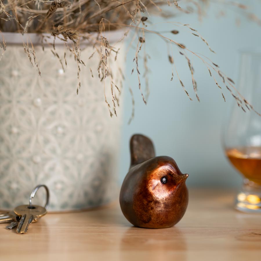 A small bronze robin sculpture on a wooden table next to a set of keys, a glass and a pot with dried flowers