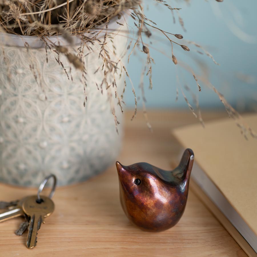 Small bronze wren sculpture on a wooden surface next to a book and some keys with a vase full of dried grasses in the background