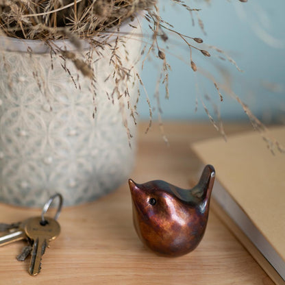 Small bronze wren sculpture on a wooden surface next to a book and some keys with a vase full of dried grasses in the background