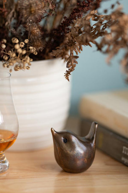 Small bronze wren sculpture on a wooden surface next to a glass and books with a white vase with dried flowers in the background