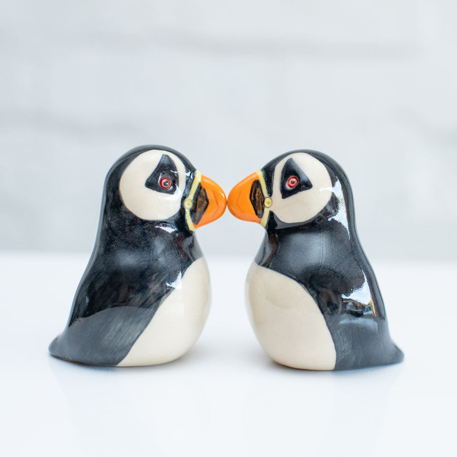 Two glossy ceramic puffin figurines facing each other, with black and white bodies and bright orange beaks, set against a clean white background.
