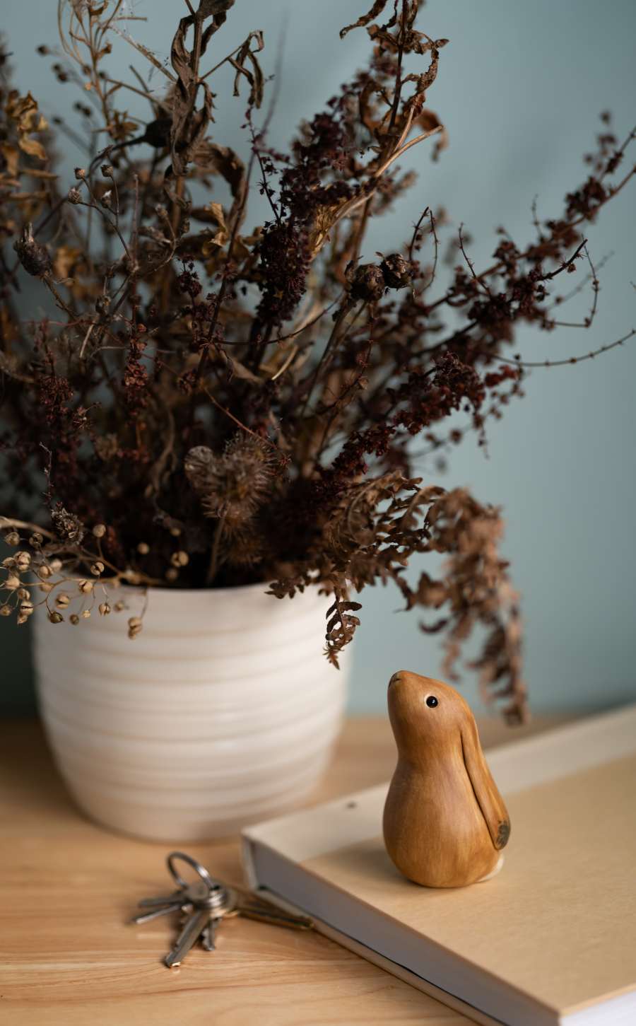 Wooden rabbit figurine on a book next to a white pot with dried plants and a set of keys