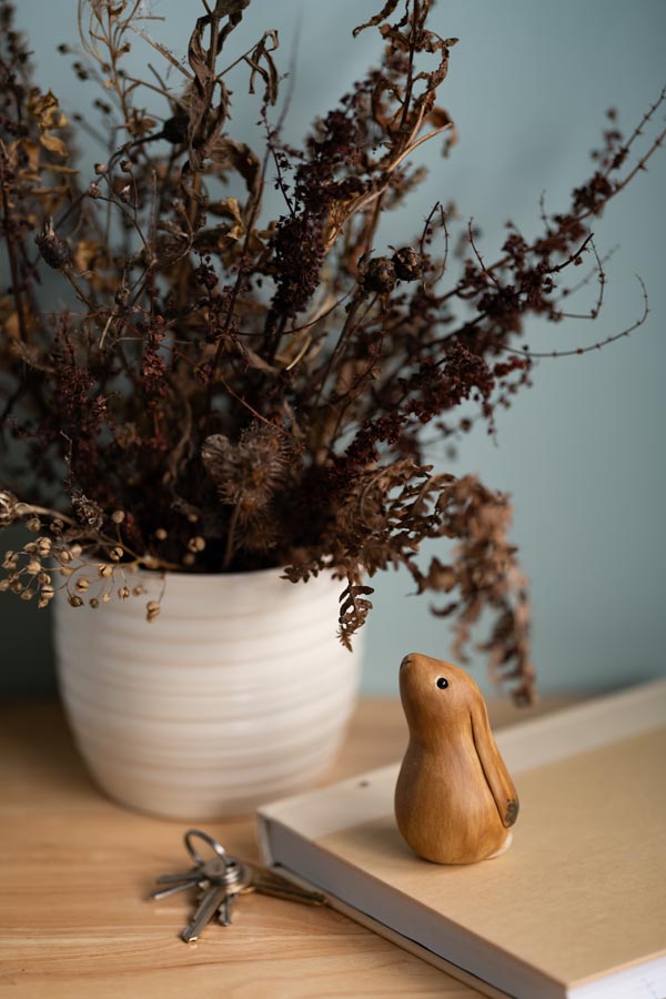 Small ceramic brown hare sculpture on a book next to some keys with a vase of dried flowers in the background