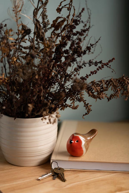 A small ceramic Robin on a book next to some keys and a white vase with dried flowers